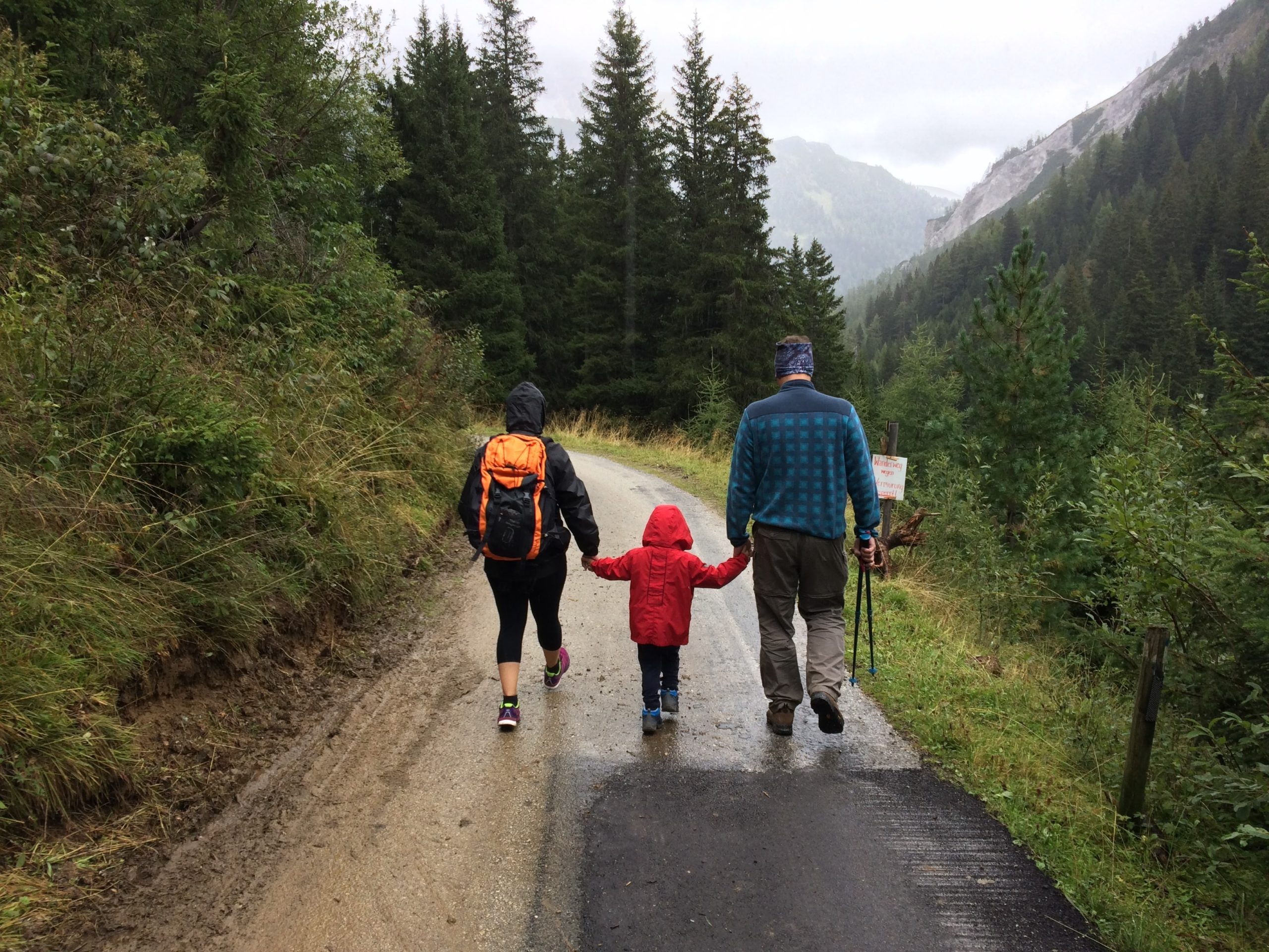 Двое родителей гуляют под дождем, а между ними ребенок. На ребенке ярко-красный плащ, и все держатся за руки. Прогулка по горной дороге