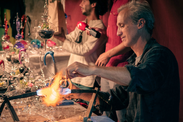 At multiple tables, glass artists use fire and various instruments to manipulate the material