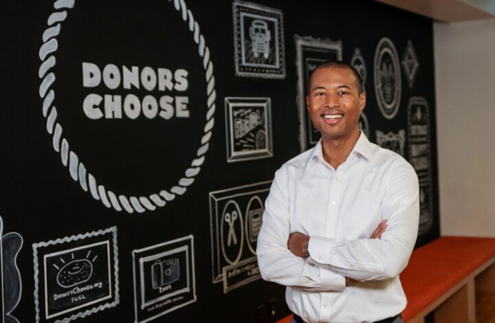 A photo of Alix Guerrier, CEO of DonorsChoose, standing in front of the DonorsChoose logo