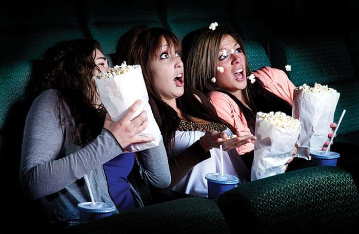 Drie mensen in bioscoopstoelen, met popcorn en drankjes, hebben een verbaasde of bange blik op hun gezicht