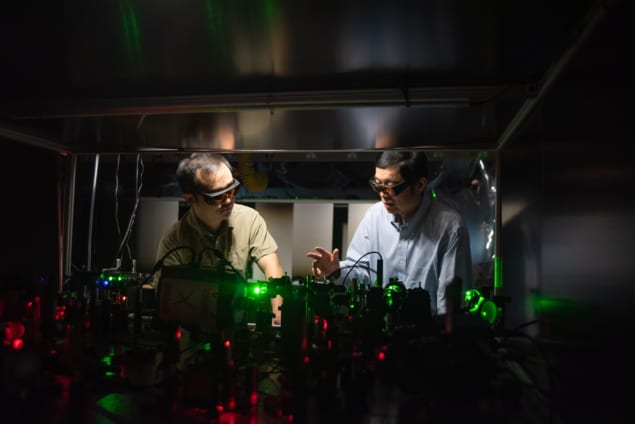 Photo of Zhe He and Lihong Wang in the lab. Both men wear laser safety goggles, and bright splotches of red and green laser light illuminate optics in an otherwise darkened foreground