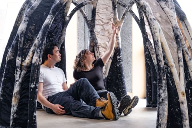 Photo of the Bioknit prototype structure, which is dome-shaped and formed of delicate, interlocking arches that are the same black, powdery colour as fungal blooms. Two people are sitting inside the structure.