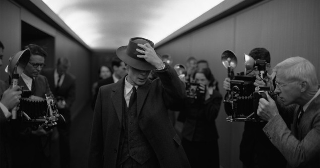 Black and white still from the film Oppenheimer showing Cillian Murphy in a corridor surrounded by press