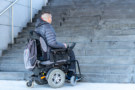 Seorang pengguna kursi roda elektrik melihat ke tangga luar. Tidak ada tanjakan atau akses alternatif lain yang terlihat