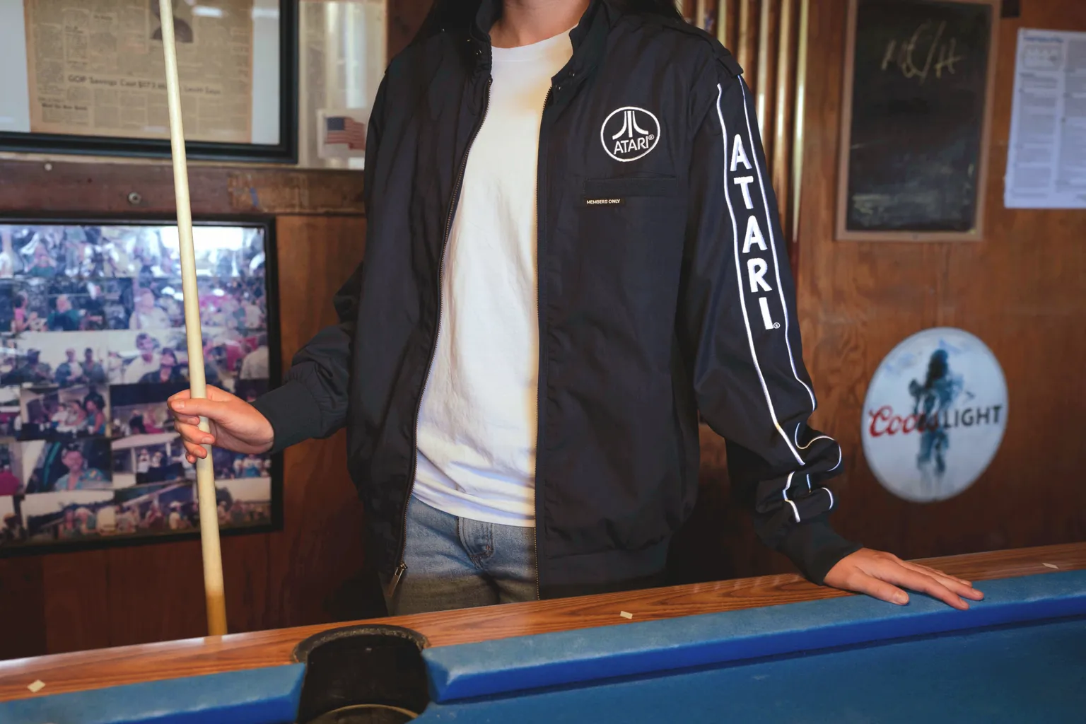 Fotografia de um homem do pescoço para baixo vestindo uma jaqueta Atari azul-marinho e prata aberta com uma camiseta branca por baixo. Ele segura um taco de sinuca e tem uma das mãos apoiada em uma mesa de sinuca azul.