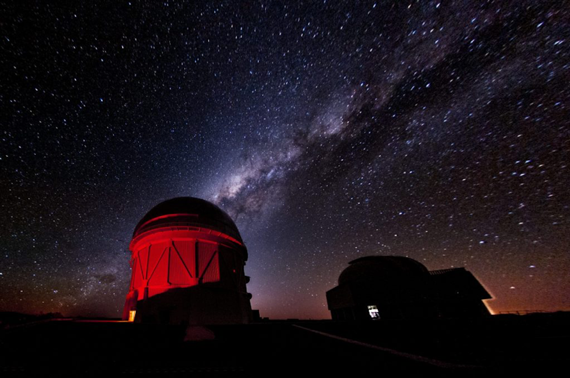 Ein Foto eines rot beleuchteten Observatoriumsgebäudes mit dem Sternenhimmel im Hintergrund.