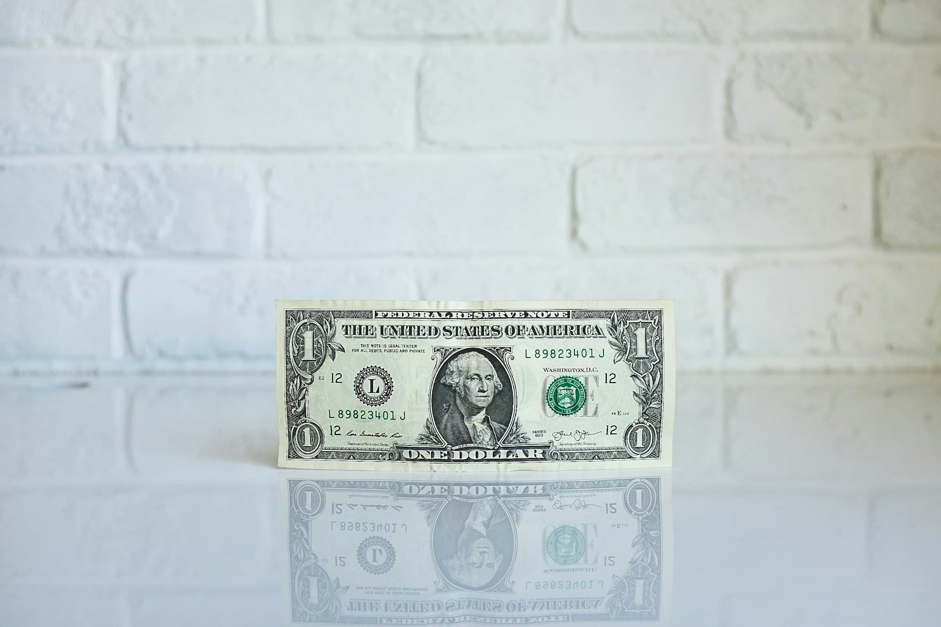 एक सफेद ईंट की दीवार की पृष्ठभूमि के सामने, जॉर्ज वाशिंगटन के चेहरे वाला एक डॉलर का बिल